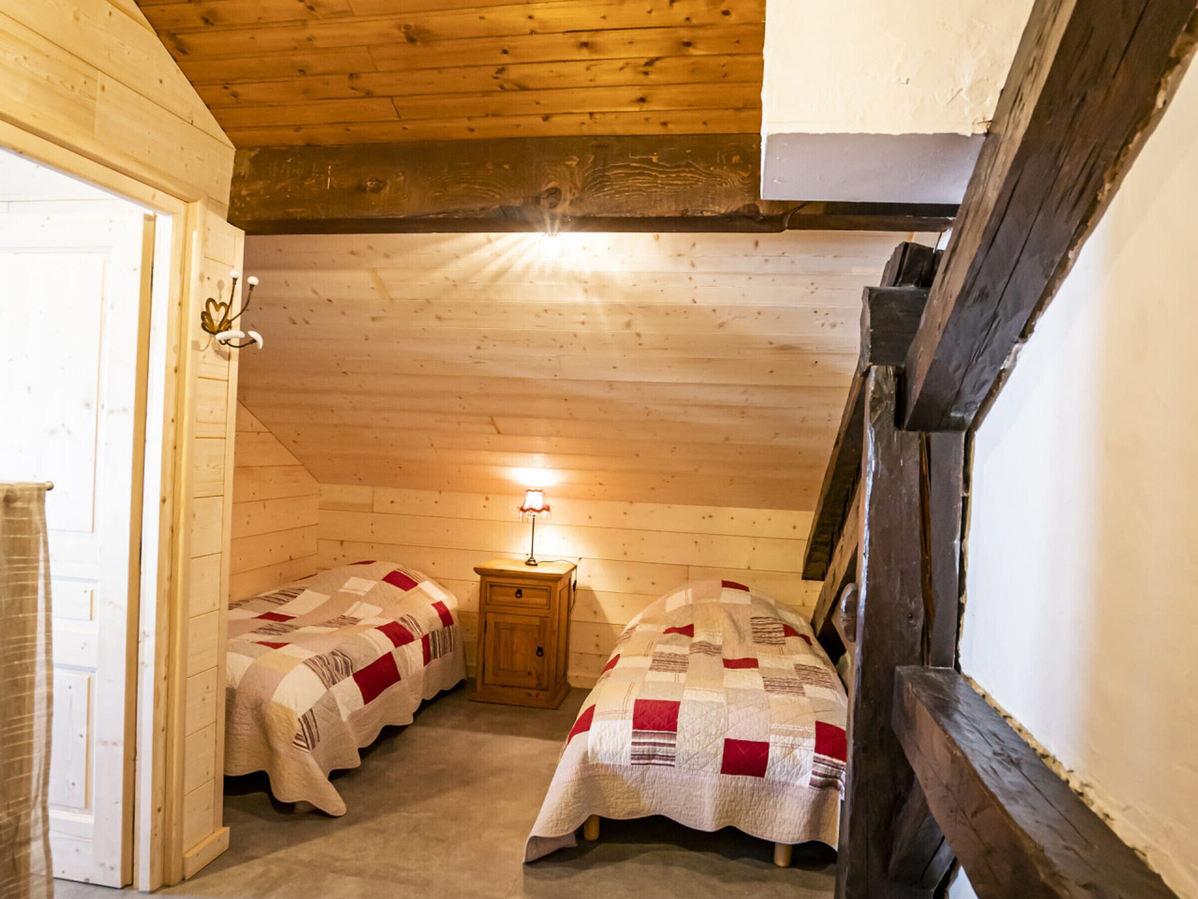 A small, cozy room with two beds, wooden walls, a sloped ceiling, and a small table with a lamp.