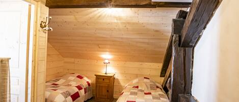 A small, cozy room with two beds, wooden walls, a sloped ceiling, and a small table with a lamp.