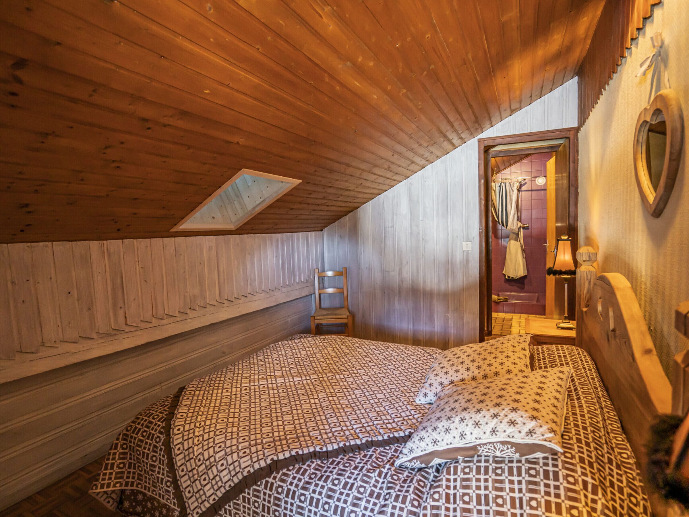 Une petite pièce chaleureuse aux murs en bois et au plafond incliné. On y trouve un lit avec une literie à motifs, une chaise et une petite table avec une lampe.