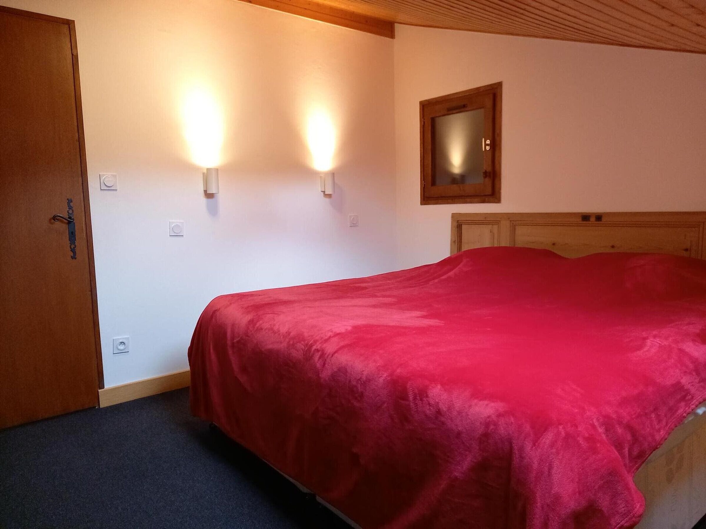 A bedroom with a bed, a wooden door, two wall-mounted lights, a mirror, and a carpeted floor.