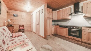 Een knus kamer in chaletstijl met houten plafond, een kitchenette met houten kastjes, een eethoek met tafel en stoelen, en een gestreept sofa.
