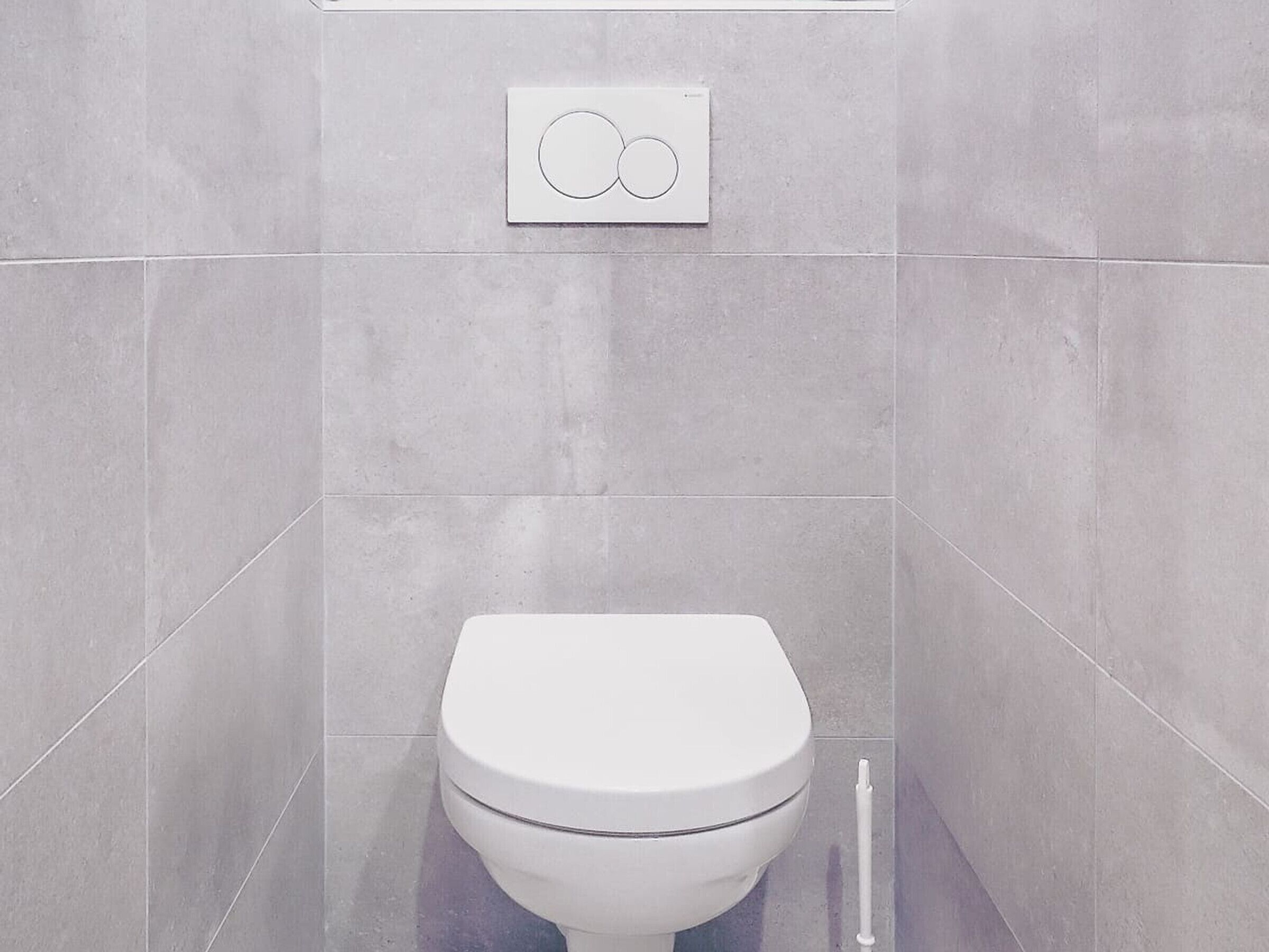A modern bathroom with a white toilet, grey tiles, and a wall-mounted flush control panel.