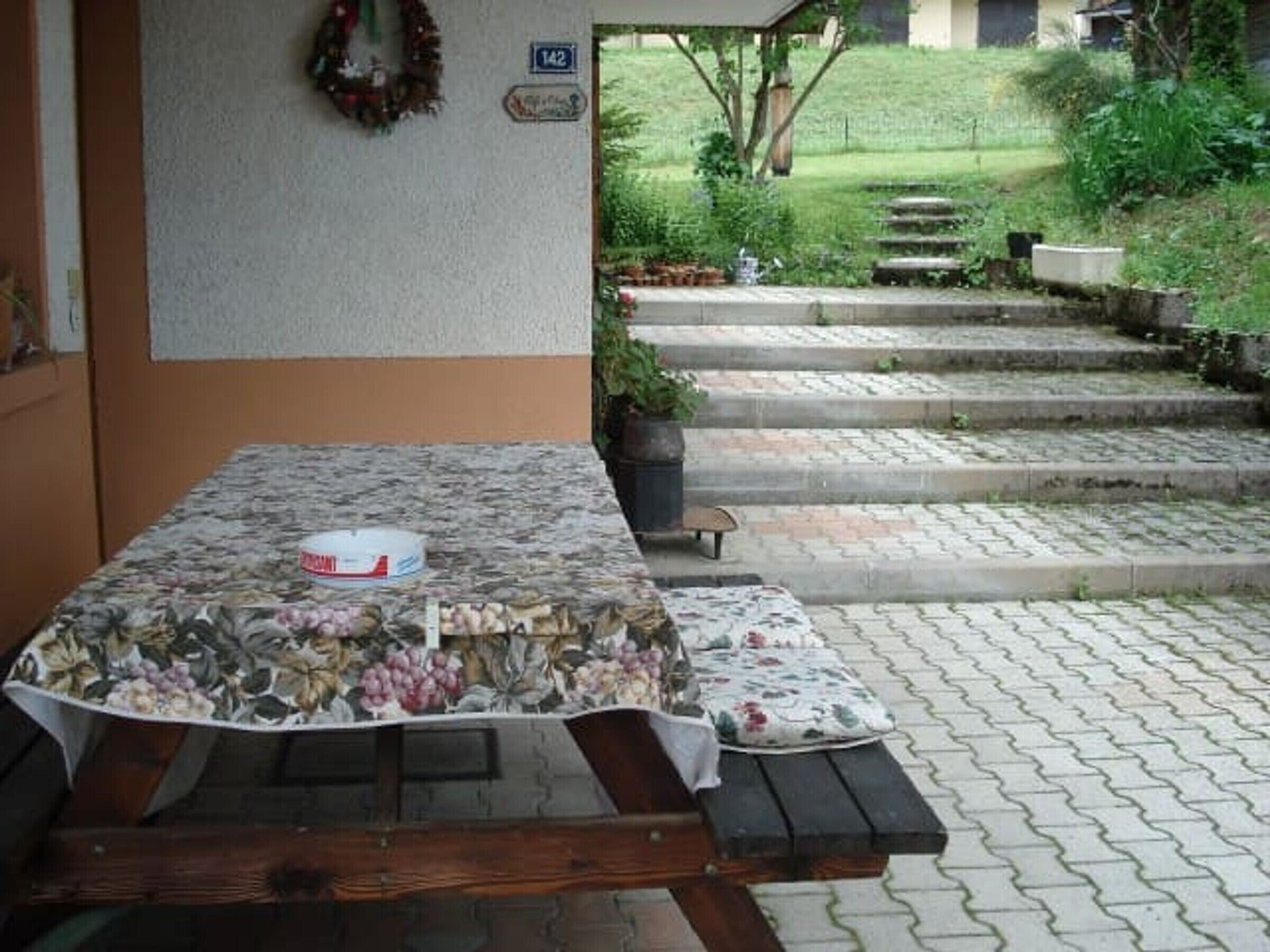 A patio with a table covered in a floral tablecloth, a bench, and a garden view.