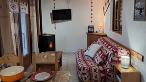 Una sala de estar acogedora, con techo de madera, un televisor en la pared, un sofá con cojines estampados, una mesa con un bol y platos, y una lámpara pequeña sobre una mesita.