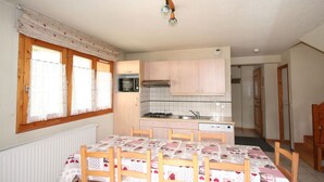Een kleine keuken met een magnetron, fornuis en spoelbak. Er is een eettafel met stoelen en een raam met gordijnen.