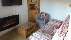A living room with a fireplace, a television, a wooden cabinet, a blue armchair, and a patterned sofa. - 2-room apt for 5 people in Châtel with south-facing balcony (Châtel)