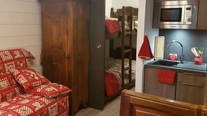 A compact room with a bunk bed, a wooden wardrobe, a kitchenette with a microwave, and a red patterned sofa.