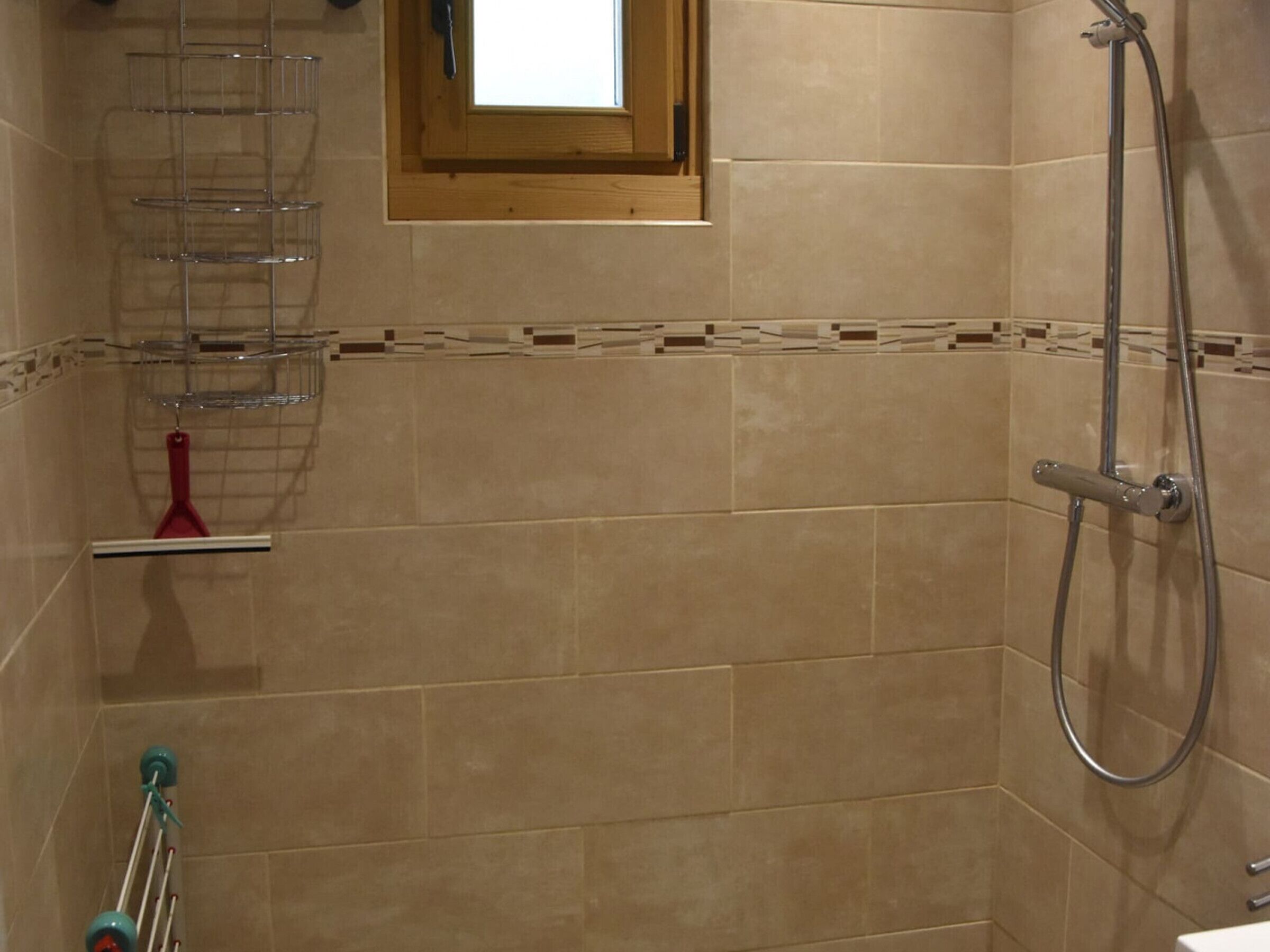 Un baño con baldosas color beige, una ducha con cabezal de ducha de mano, una ventana con marco de madera y un perchero con una toalla roja.
