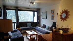 A cozy room with a bed, a sofa, a desk, and a dining table. There are large windows with a view of trees and a building outside.