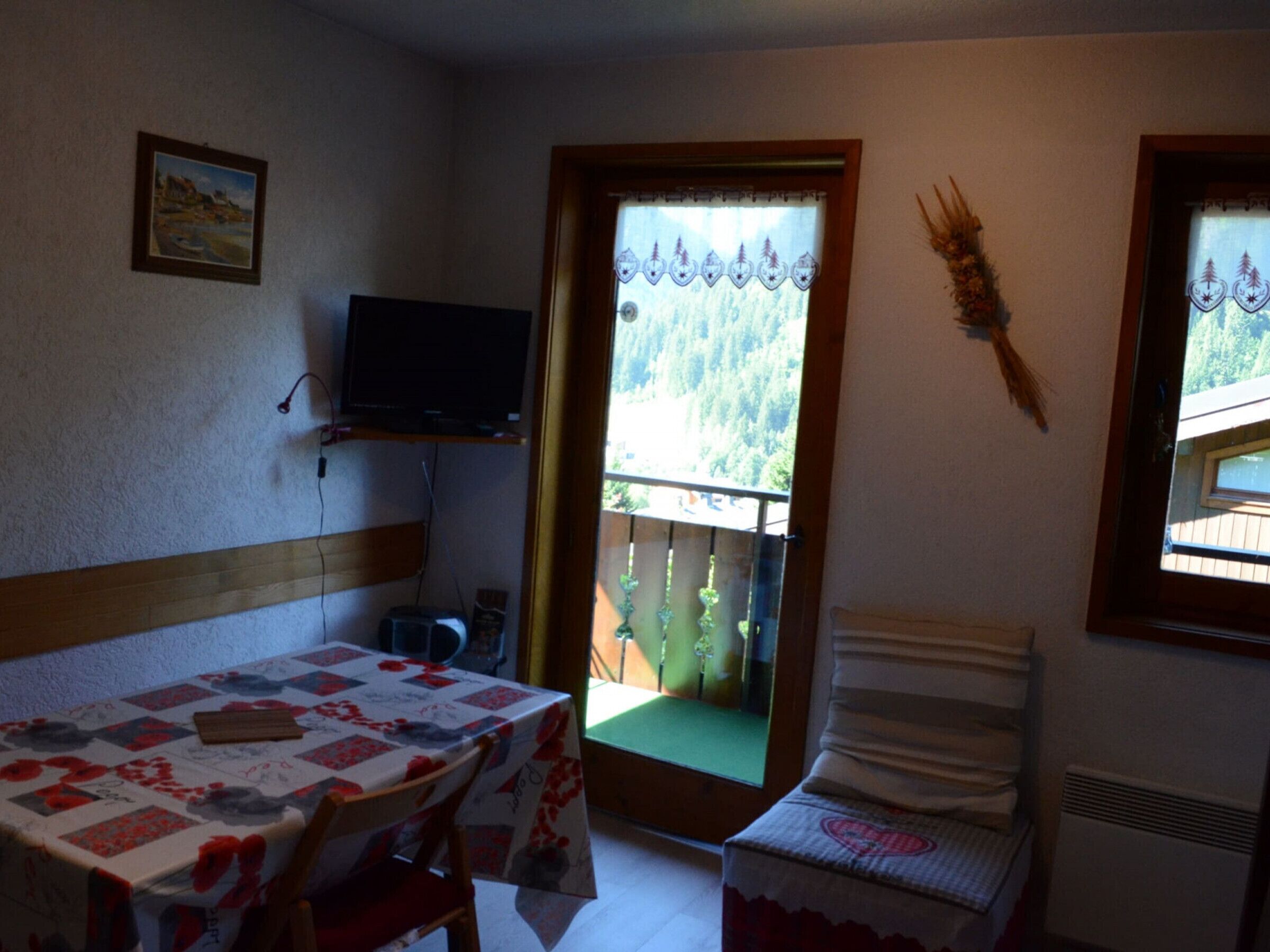 Une petite pièce avec une table recouverte d’une nappe à motifs floraux, une chaise, un téléviseur posé sur une étagère et une fenêtre donnant sur une forêt.