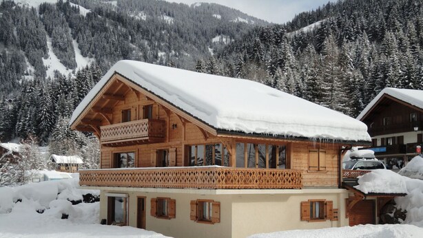 Una cabaña de madera con balcón, rodeada de árboles nevados y montañas.
