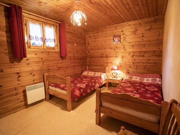 A cabin-style bedroom with two wooden beds, a window with curtains, a heater, and a hanging light fixture.