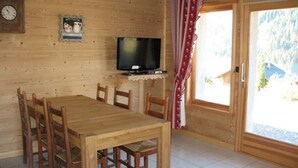 Comedor de madera con una mesa, sillas, televisor sobre un mueble, un reloj en la pared y una puerta que ofrece vistas al exterior.