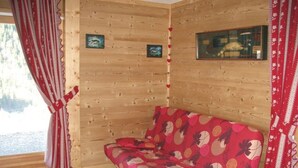 Interior de una cabaña de madera con un sofá rojo estampado, una ventana con cortinas rojas y dos cuadros en las paredes.