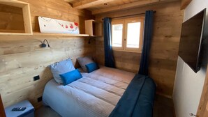 Tunnelmallinen mökkimaja, jossa on sänky, pieni pöytä, koriste-esineillä varustettu hylly ja ikkuna, jossa on verhot.