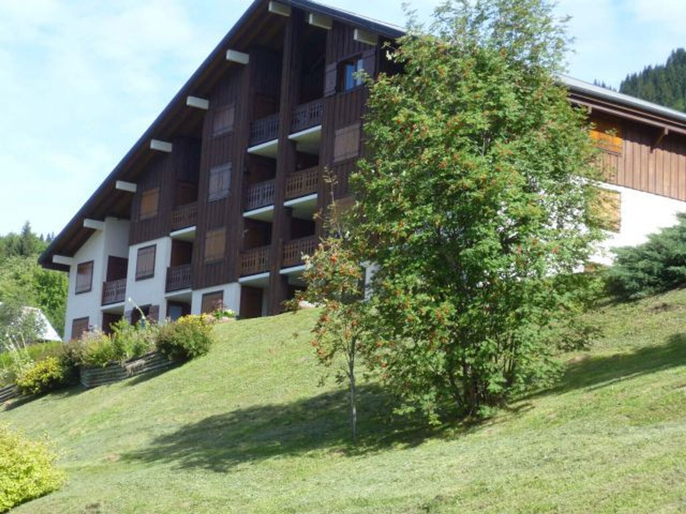 A multi-story building with balconies, surrounded by greenery and a sloped landscape.