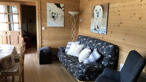 A cozy wooden cabin interior with a wooden dining table, a sofa with patterned cushions, and two paintings of polar bears on the walls. - Cozy 2BR Apartment, South-Facing Terrace, Ski Access, WiFi, Parking, 4 Guests (Châtel)