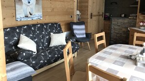 A cozy living room with a wooden wall, a sofa with patterned cushions, a chair, and a dining table with a checkered tablecloth. - Cozy 2BR Apartment, South-Facing Terrace, Ski Access, WiFi, Parking, 4 Guests (Châtel)