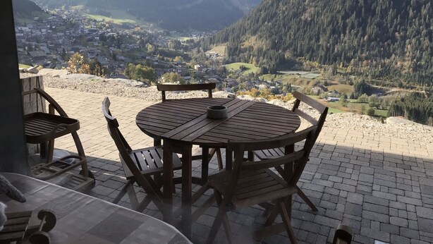A wooden table and chairs on a patio with a view of a valley and mountains. - Cozy 2BR Apartment, South-Facing Terrace, Ski Access, WiFi, Parking, 4 Guests (Châtel)