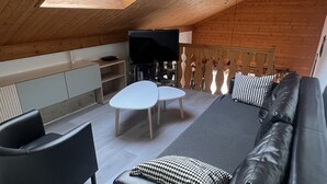 Moderni, avoimen pohjaratkaisun asunto, jossa on korkea katto, puupalkit, sohva, sohvapöytä, televisio ja parveke, jossa on puinen kaide.