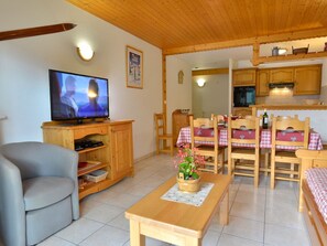 A cozy living room with a television, wooden furniture, a dining table with chairs, and a kitchen area.
