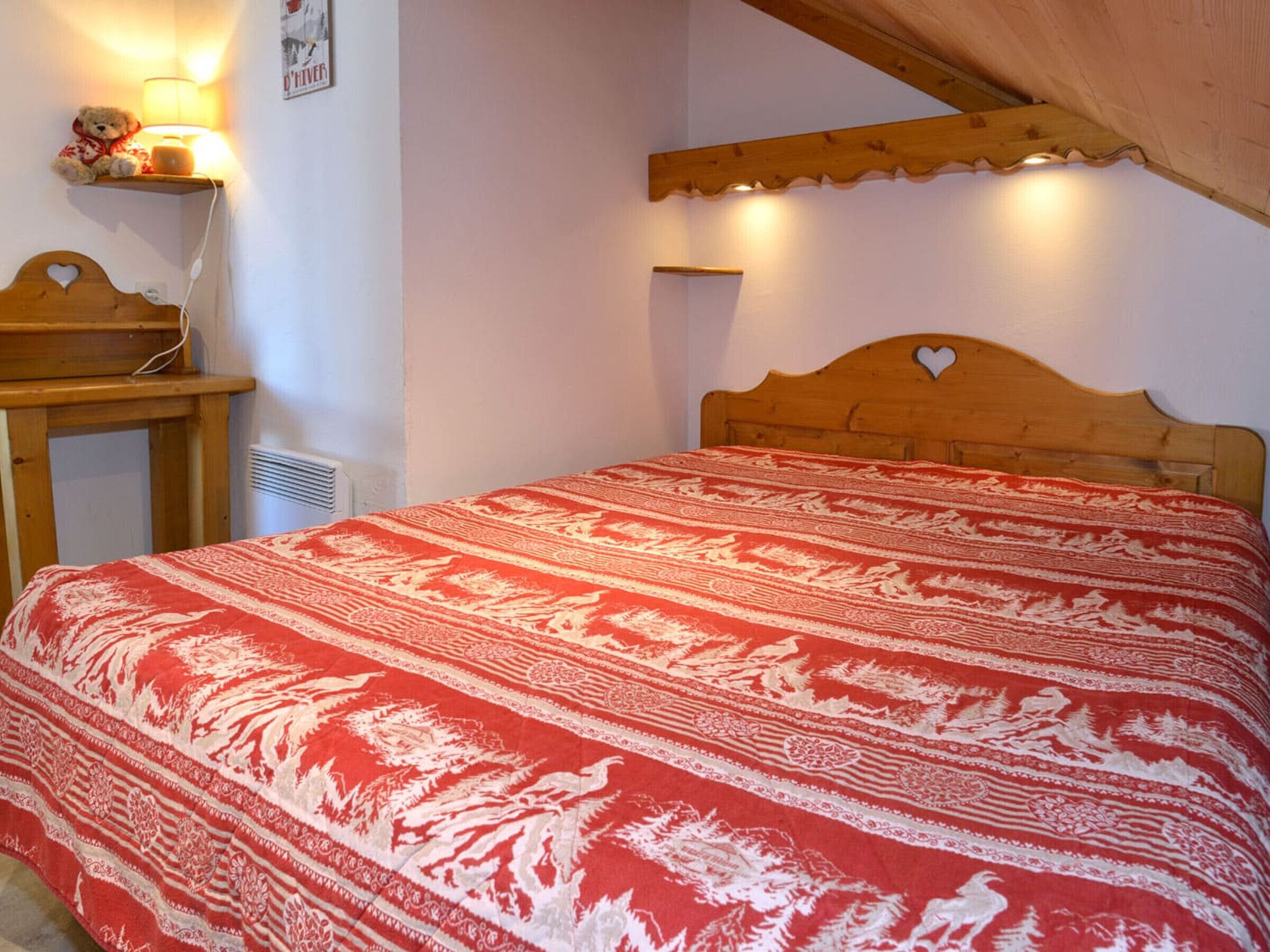 Un dormitorio con estructura de cama de madera, colcha estampada en rojo y blanco, mesita de noche con lámpara y estante montado en la pared con un oso de peluche.