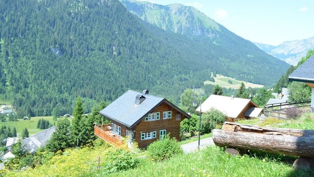 Ett trähus med ett sluttande tak, omgivet av frodig grönska och med berg i bakgrunden.