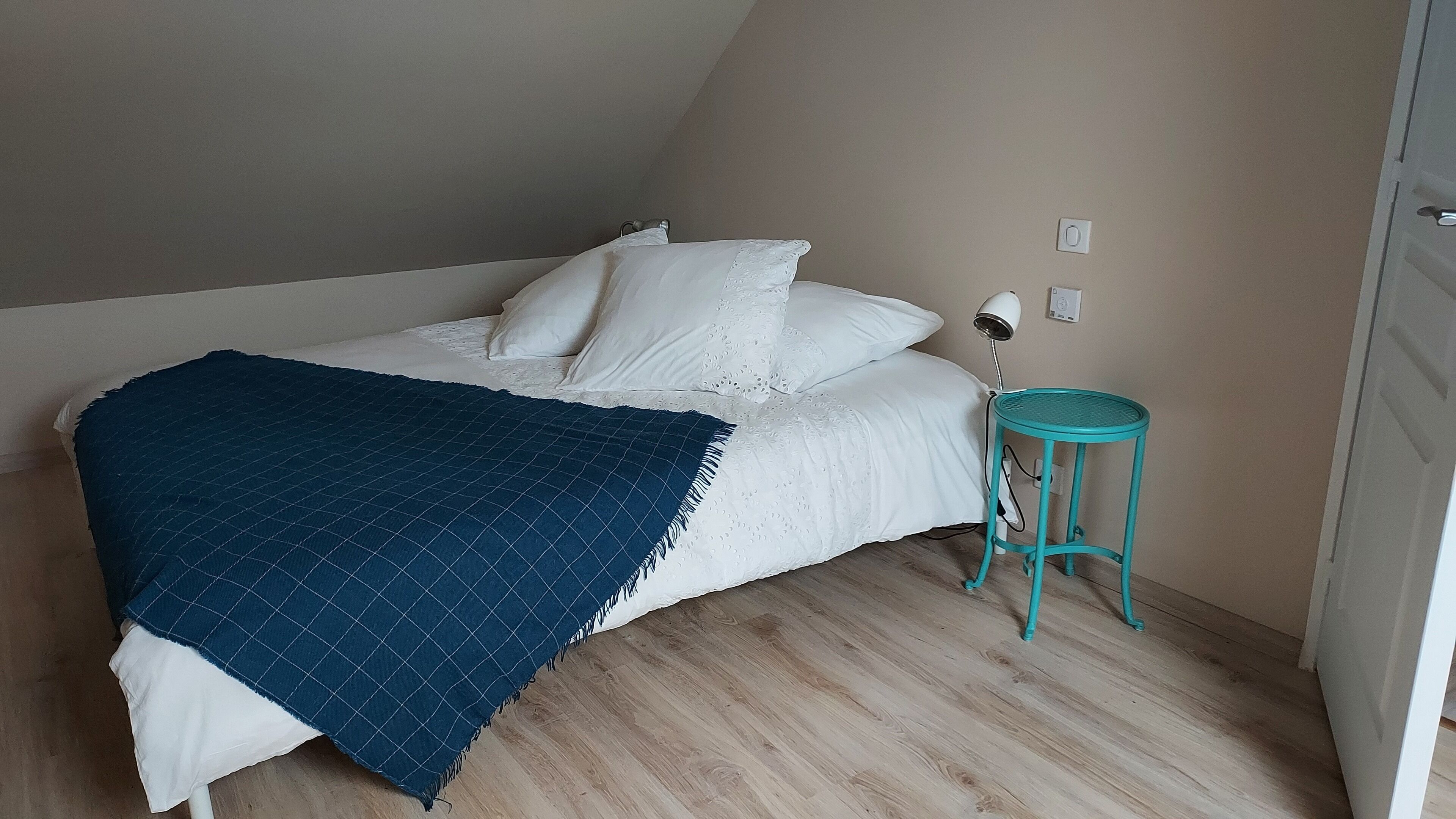 A bedroom with a bed, a blue blanket, a white pillow, a small teal stool, and a bedside lamp.