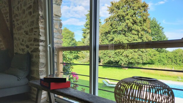 Una habitación con un ventanal grande, un banco de madera, una lámpara de mimbre y vistas a una piscina y jardín.