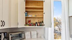 Cocina compacta con armarios blancos, microondas integrado y una ventana con cortinas.