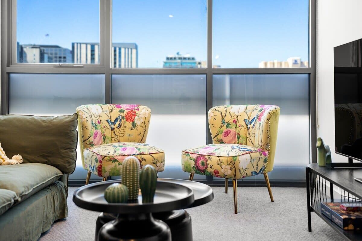 A modern living room with a large window, two floral armchairs, a sofa, a coffee table with a cactus, and a TV on a stand.