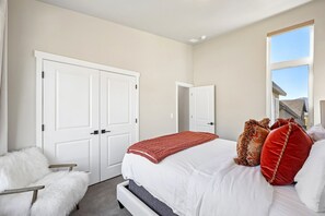 A bedroom with a large bed, a white fluffy chair, and a window with a view of a house and mountains. - Deer Springs 4BR | Private Hot Tub | Deer Valley & Jordanelle Views (Hideout)