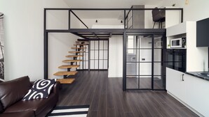 Ein modernes Interieur mit einer Treppe, die zu einem Schlafzimmer auf dem Dachboden führt, einer Küchenzeile und einer Sitzecke mit einer Couch und einem Kissen mit Muster.