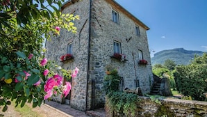 A stone building with multiple windows and flower boxes, surrounded by greenery and a mountainous backdrop. - Farmhouse with Farm in the hills of Barga: Casa Chioi (barga)