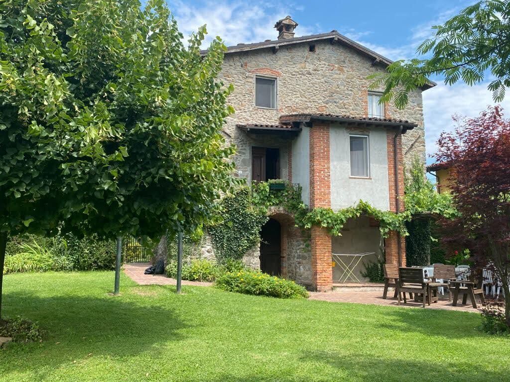 Un edificio de piedra con base de ladrillo, una ventana pequeña y una puerta. Frente, un área con césped, una banca y una mesa.