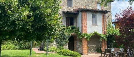 Un edificio de piedra con base de ladrillo, una ventana pequeña y una puerta. Frente, un área con césped, una banca y una mesa.