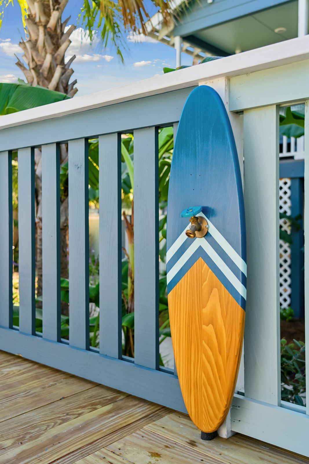 A surfboard leaning against a white railing with a blue and yellow design, set against a backdrop of greenery and a clear sky.