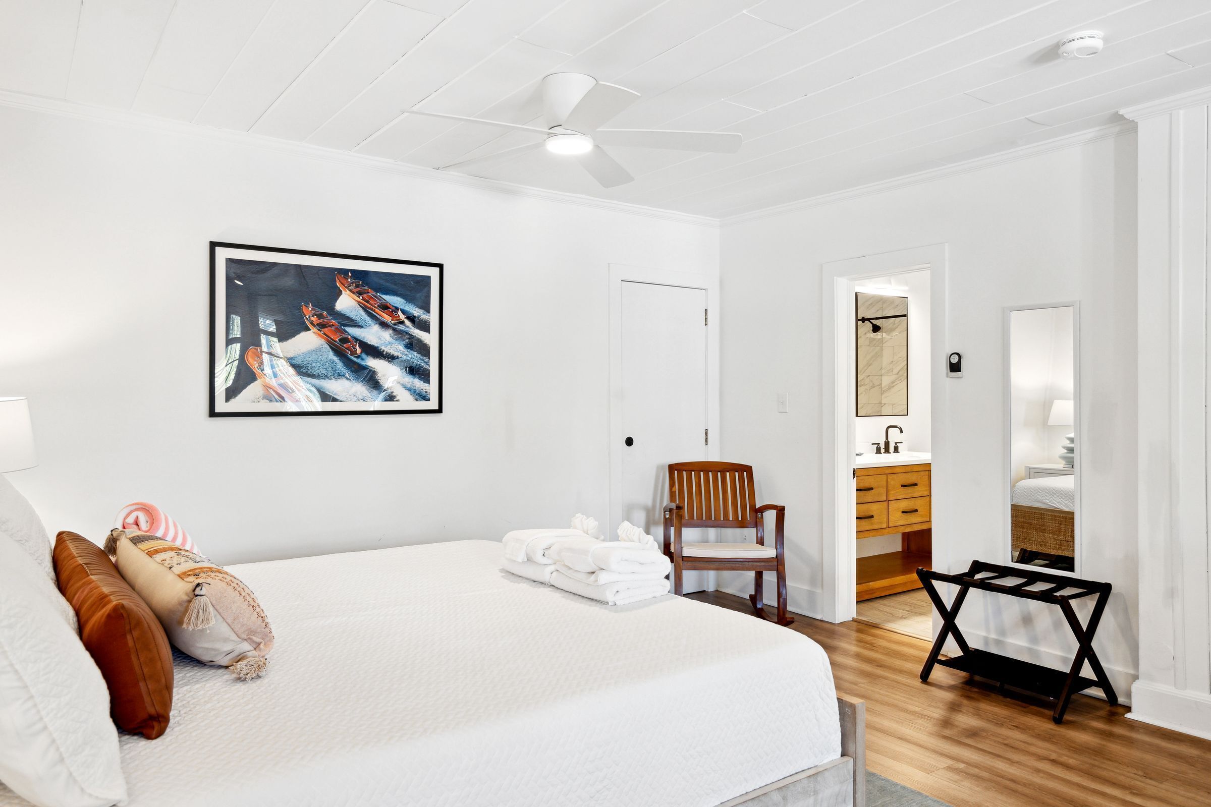 A hotel room with a large bed, a ceiling fan, a painting of boats, a wooden chair, a small table, and a bathroom visible through an open door.