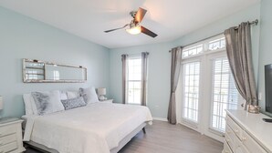 A bedroom with a bed, a ceiling fan, a dresser, a TV, and large windows with curtains. - Seaside at Anastasia E305 - Stunning beach rental with great location (St Augustine)