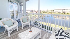 A covered balcony with wicker furniture, a view of a water body, and a row of buildings in the background. - Seaside at Anastasia E305 - Stunning beach rental with great location (St Augustine)