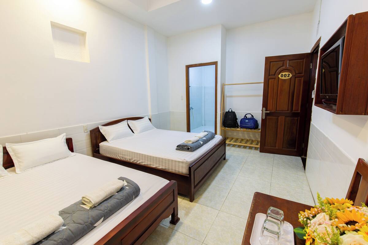 A hotel room with two single beds, a wooden wardrobe, a small table with a vase of flowers, and a door marked with the number 002.