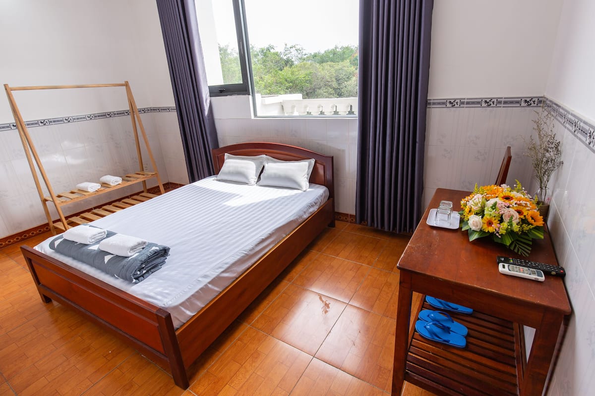 A neatly made bed with a wooden frame and white linens, a wooden bedside table with a vase of flowers and a remote control, and a window with curtains.