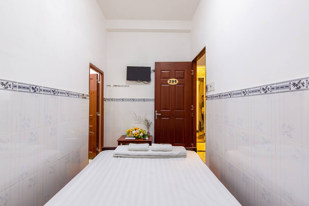 A hotel room with a bed, a wooden door with the number 204, a small table with a vase of flowers, and a television mounted on the wall.