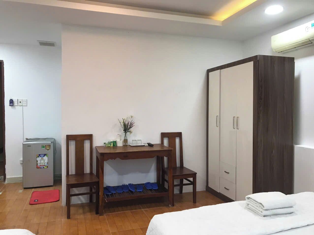A hotel room with a bed, a wooden table with chairs, a small refrigerator, and a wardrobe.