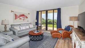 Un salón amplio con un sofá gris, una mesa de centro redonda y una silla de mimbre. Un cuadro grande de peces decora la pared, y un televisor está montado allí.