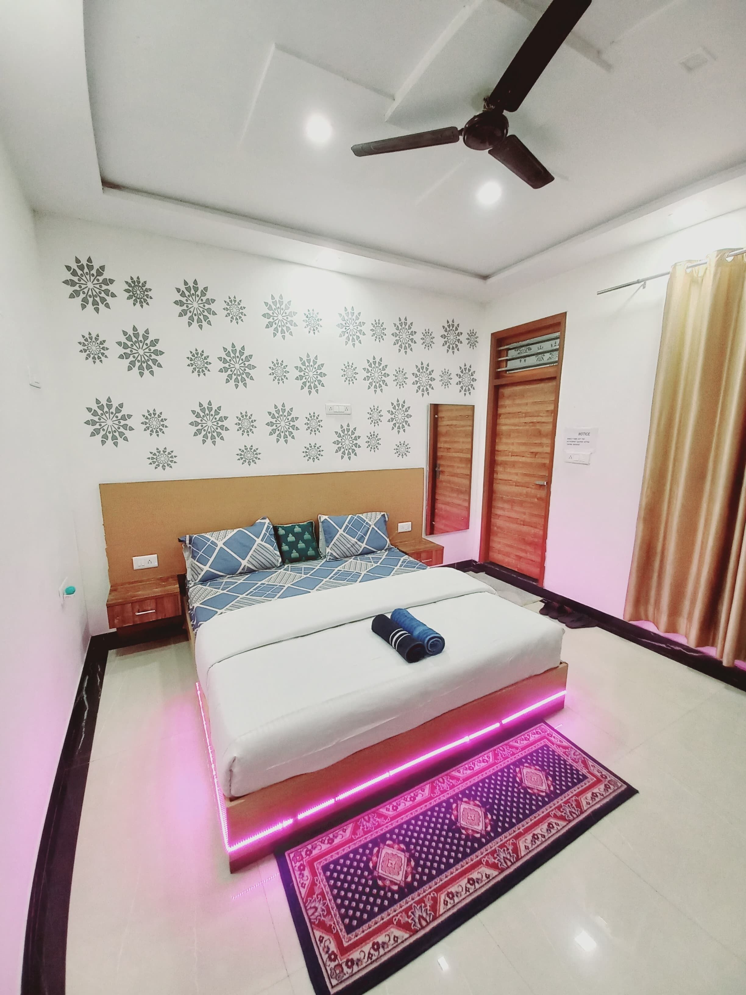A bedroom with a bed, a ceiling fan, a patterned wall, a wooden door, and a patterned rug.