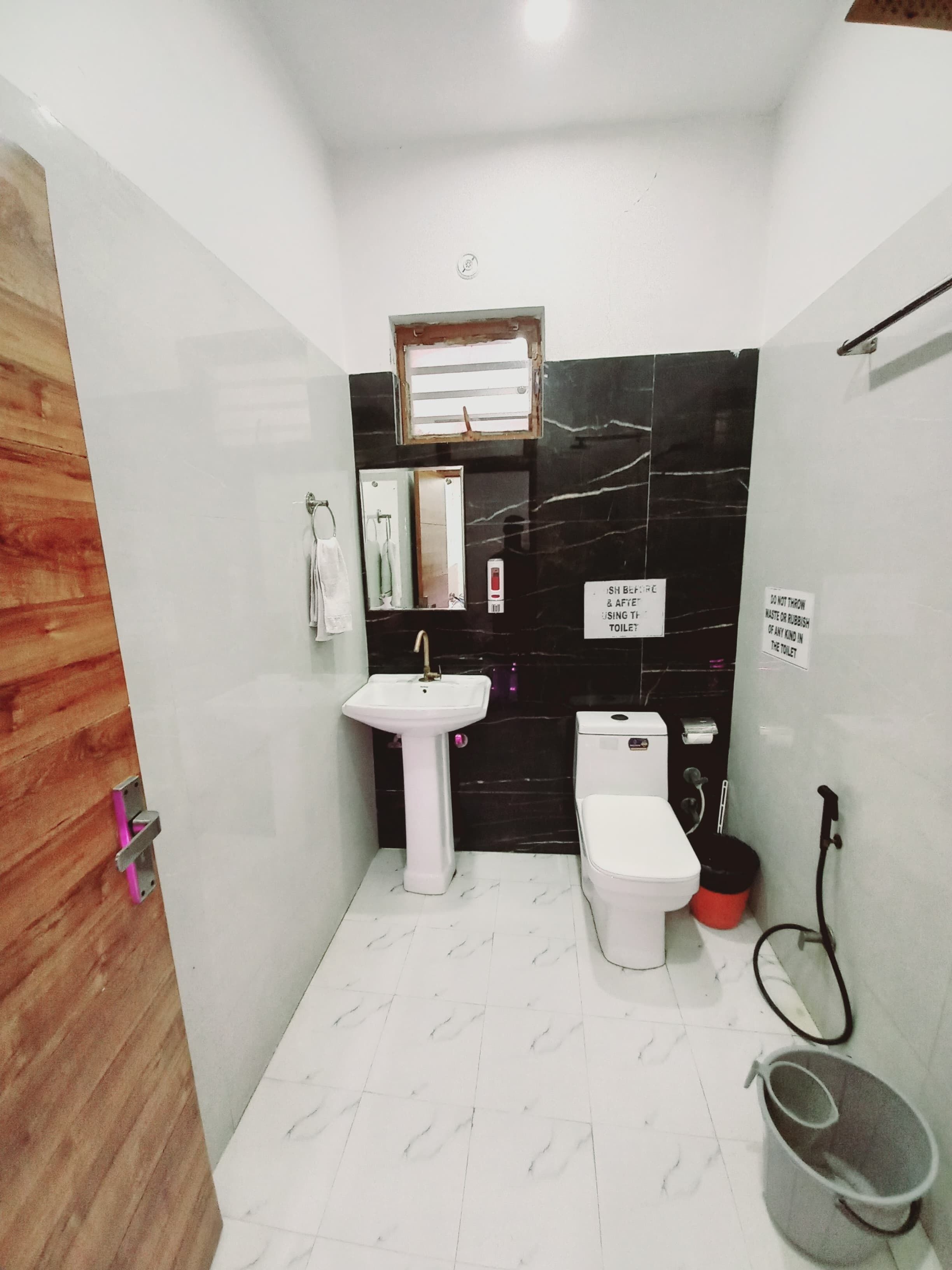 Un baño pequeño y moderno con lavamanos, inodoro y ducha blancos. Una puerta de madera se encuentra a la izquierda y una pared con azulejos negros. Un letrero en la pared indica no arrojar residuos ni basura en el inodoro.