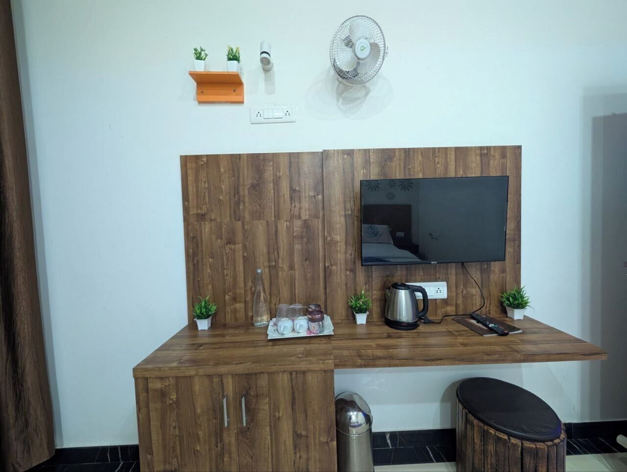 A compact living space with a wooden desk, a flat-screen TV, a small stool, and a wall-mounted fan.