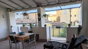 A covered patio with a wooden table, chairs, and a cushioned seating area. There are potted plants and a view of buildings in the background. - Lovely space with terrace in the old town! (Catania)