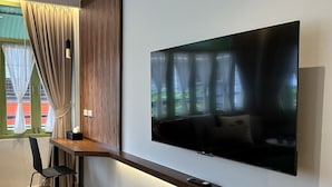 A modern living room with a large flat-screen TV mounted on the wall, a wooden desk with a chair, and a view of greenery through the window. - The Tangerine Guesthouse (Bangkok)
